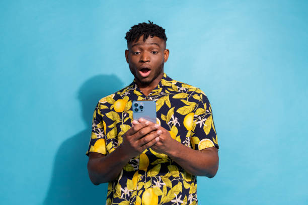 istock Young man in stylish floral shirt using smartphone with surprised expression on blue background 2191291715