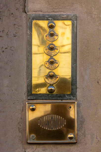 Polished brass doorbell panel mounted on a stone wall. The panel features multiple buttons arranged in a vertical line, each likely corresponding to a different apartment or office. Below the buttons is a brass speaker grille, part of the intercom system. The elegant design and craftsmanship suggest it belongs to a historic or upscale building, possibly in a European city like Florence.