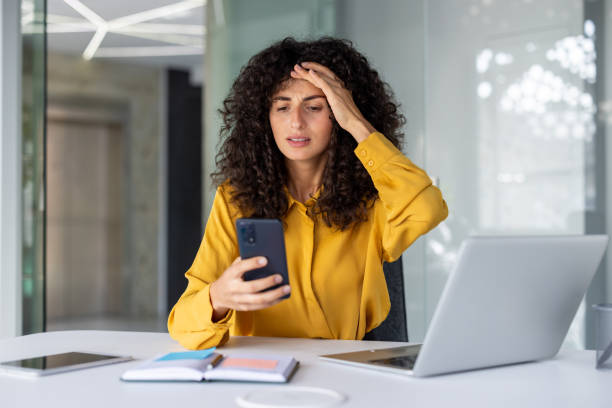 istock Young woman in yellow blouse looking concerned at smartphone in modern office while working on laptop and notebook 2191294361