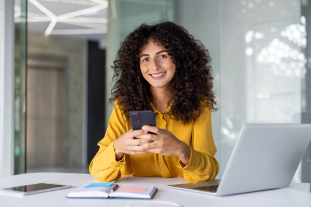 istock Smiling woman using smartphone in modern office setting with laptop and open notebook, showcasing technology and communication 2191296396