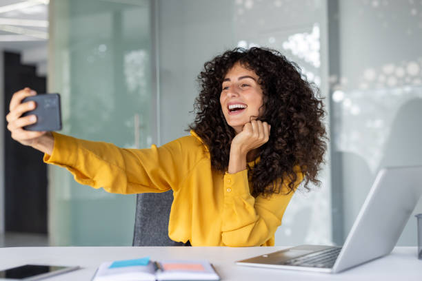 istock Young woman in vibrant office setting capturing selfie using smartphone expressing joy with bright smile. Professional atmosphere highlighted by laptop and organized workspace accessories. 2191300313