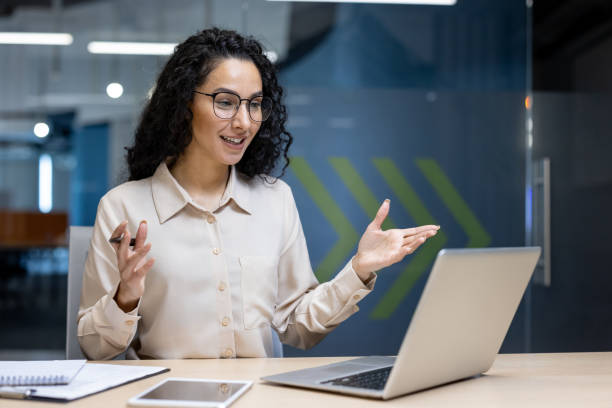istock Smiling professional woman in office on video call using laptop computer, gesturing enthusiastically, with smartphone and notebook on table, wearing glasses and white shirt 2191300318