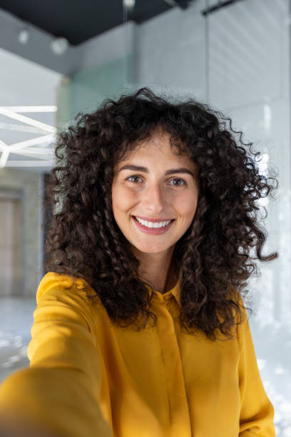Smiling woman taking a selfie indoors with a smartphone, showcasing a joyful expression and curly hair. She is wearing a bright yellow shirt, standing in a modern, well-lit room.