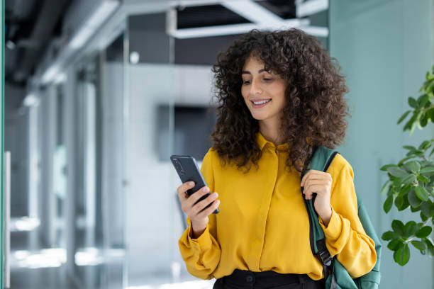 Smiling young student with curly hair using smartphone while walking through modern college hallway