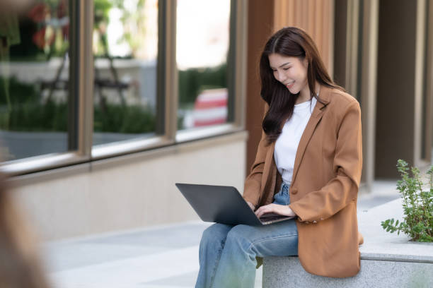 istock woman in brown blazer sits outside, working on laptop with smile. modern setting features greenery and large windows, creating relaxed atmosphere for productivity 2191306093