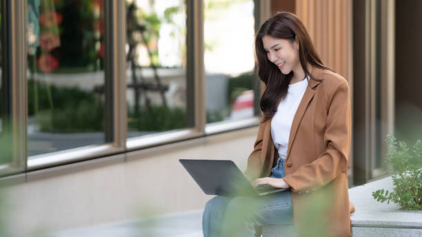 istock woman sits outdoors, smiling while using laptop. She wears brown blazer and jeans, surrounded by greenery and modern architecture, creating relaxed atmosphere 2191306095
