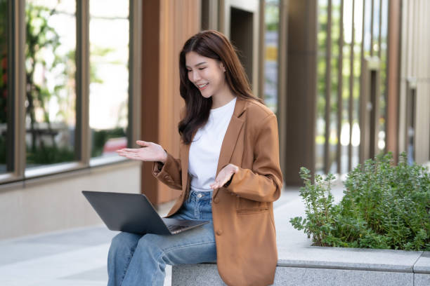 istock young woman in brown blazer sits outdoors, smiling while using laptop. She appears engaged and happy, surrounded by greenery and modern architecture 2191306103