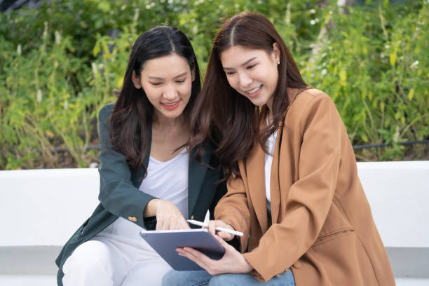 Two women collaborating on tablet outdoors, sharing ideas and smiles. scene captures moment of teamwork and creativity in vibrant green setting