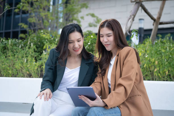 istock Two women sitting together, engaging with tablet outdoors, showcasing collaboration and technology. Their expressions reflect interest and enjoyment in moment 2191306108