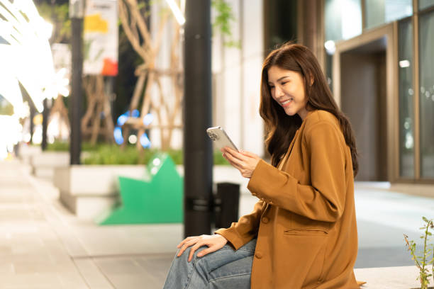istock woman smiles while using her smartphone outdoors at night, dressed in stylish brown coat. urban setting features soft lighting and greenery, creating relaxed atmosphere 2191306112