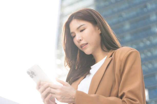 istock woman in brown blazer is using smartphone outdoors, surrounded by modern buildings. sunlight creates bright atmosphere, highlighting her focused expression 2191306122