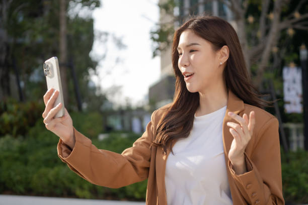 istock woman is smiling and waving while using smartphone outdoors in sunny park. Her casual outfit and joyful expression convey sense of connection and happiness 2191306130