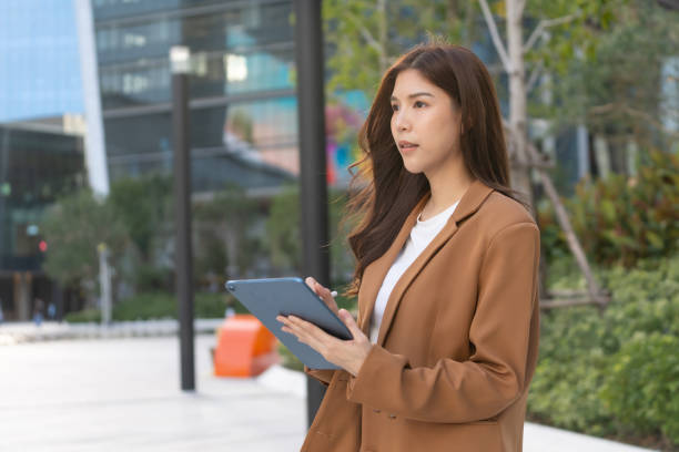istock woman in brown blazer holds tablet outdoors, showcasing modern architecture and greenery. She appears focused and engaged in her task 2191306170