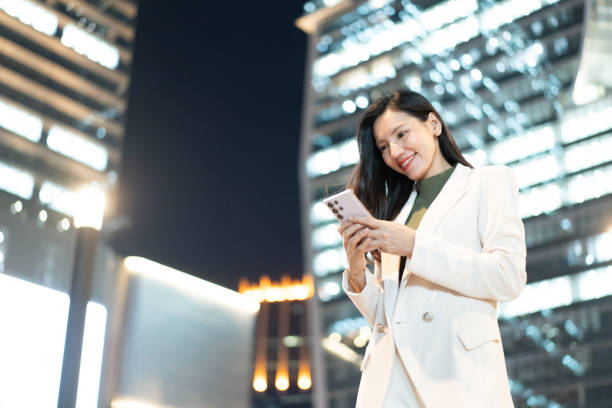 istock woman in white suit smiles while using her smartphone at night in urban setting, surrounded by modern buildings 2191306297