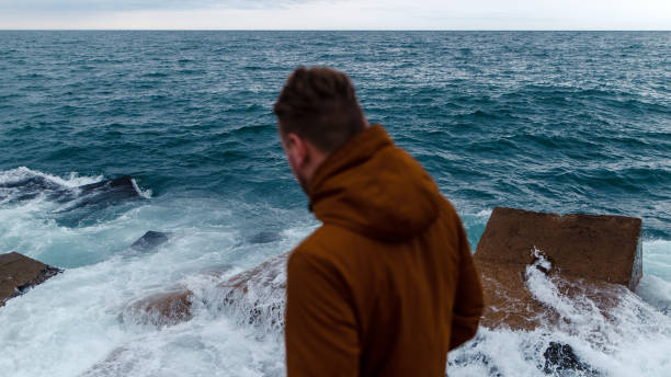 A person in a brown jacket stands on a rocky shore, looking at the ocean waves crashing against the rocks under a cloudy sky.