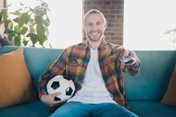 istock Young man relaxing at home on weekend holding soccer ball and remote control 2191308726