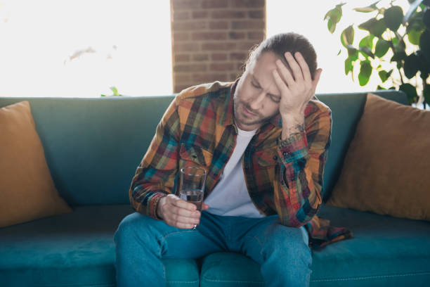 istock Young man in a checkered shirt holding a glass while sitting on a sofa, appearing thoughtful in a cozy living room setting 2191308889