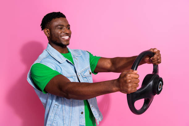 istock Young man smiling while holding a steering wheel against a vibrant pink background, showcasing casual denim fashion 2191309436