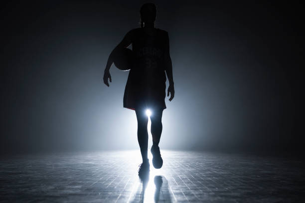 Female basketball player silhouette training at dark indoor court illuminated with lights wide shot. Team sport and healthy lifestyle concept.