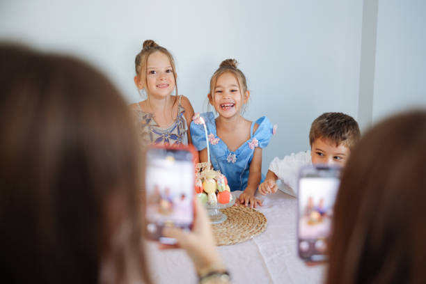 Child birthday party. Adorable little boy and girl with birthday cake decorated flowers and burn candle, makes wish. Kid in festive dress at room with balloons, bouquet of flowers at vase