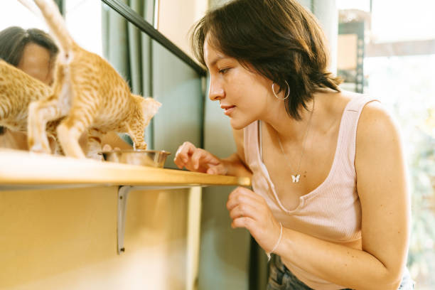 Young woman observes a small ginger kitten as it eats from a metal bowl placed on a wooden table