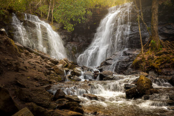 This image depicts Soco Falls in North Carolina, showcasing the waterfall cascading over rugged rocks in a serene forested setting. The sunlight filtering through the trees highlights the lush surroundings and the intricate textures of the stones.