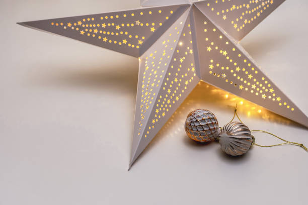 istock Glowing paper Christmas star and two Christmas balls on a white table. 2191353216