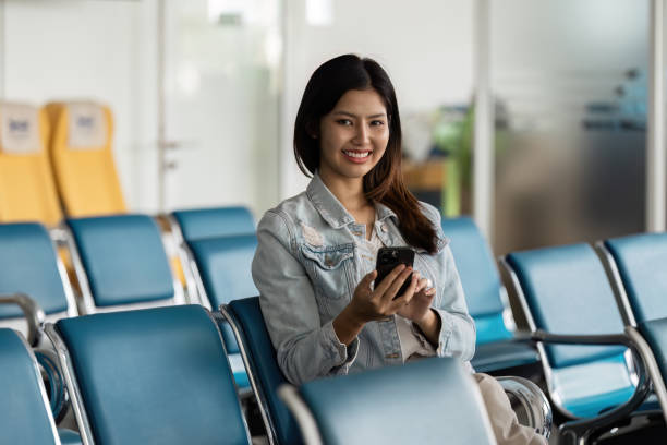 istock Young Woman Smiling While Using Smartphone in Modern Airport Waiting Area with Blue Chairs and Bright Natural Lighting 2191358008