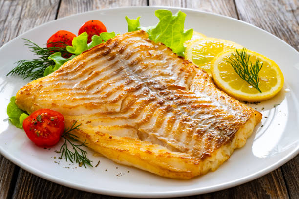 istock Fried cod fillet, boiled potatoes, fresh vegetables and lemon slices served on white plate on wooden table 2191359702