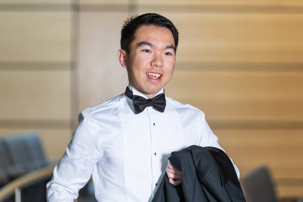 A young university student man standing in an auditorium, wearing a suit with bow tie while holding onto his tuxedo jacket, participating in a formal reception event