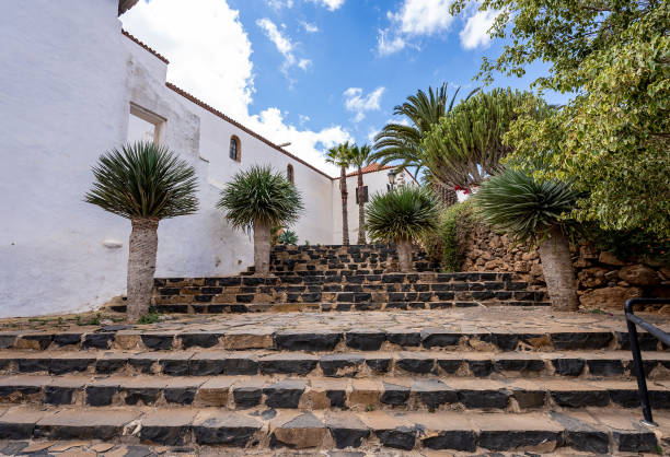Photograph of Betancuria, one of the most historic villages in Fuerteventura, Canary Islands. The image captures the stone fountain, whitewashed walls, and the church tower surrounded by flowers under a clear blue sky. A perfect representation of Canarian architecture and heritage, ideal for travel, culture, and tourism projects.