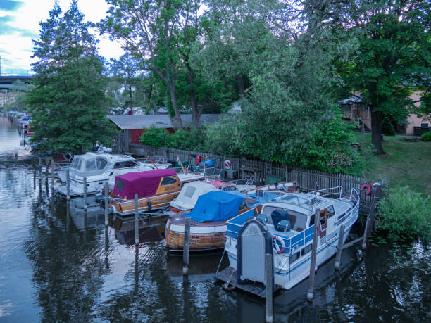 Stockholm, Sweden - 13 June 2024: A serene view of a marina with several boats docked along a calm waterway. The boats vary in size and design, surrounded by lush greenery and trees. A wooden fence and a small building are visible in the background, adding to the tranquil atmosphere.
