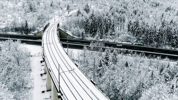 A striking aerial view of a snow-covered railway bridge crossing over a highway amidst a serene winter forest. The image highlights the integration of modern transportation infrastructure with the natural beauty of winter landscapes.