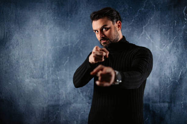 Portrait of a mid-adult man pointing at the camera while standing against a gray background.