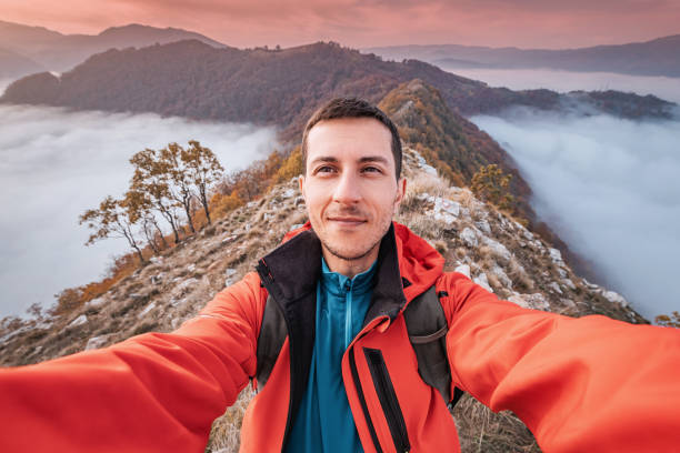 Young hiker taking a selfie on a mountain peak above the clouds during a colorful sunset