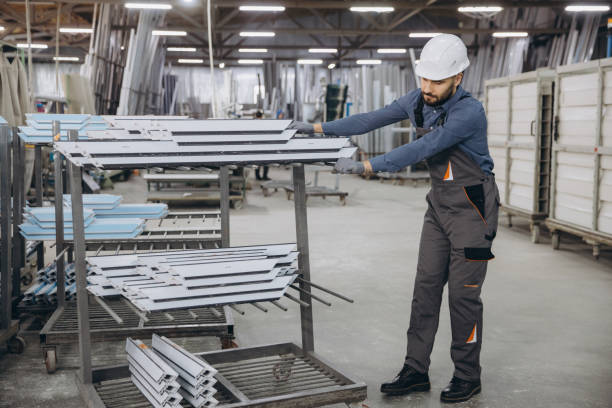 Factory worker wearing uniform and hardhat moving aluminum window profiles inside a production plant
