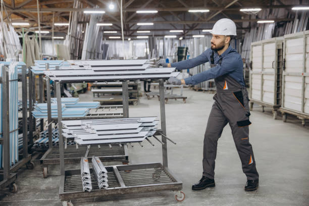 Factory worker wearing uniform and hardhat pushing cart with aluminum profiles in a factory for windows and doors production
