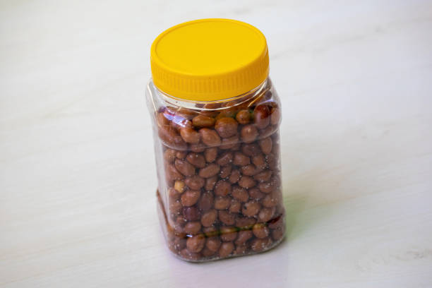 A clear glass jar filled with peeled, crunchy roasted peanuts placed on a wooden table, highlighting a tasty snack and cooking ingredient