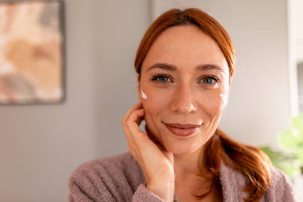 istock Smiling woman applying moisturizer cream on face looking at camera 2191394929
