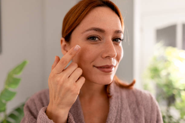 Smiling woman applying moisturizer cream on face looking at camera