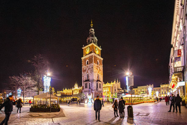 Krakow, Poland - December 5, 2018: Krakow’s Town Hall Tower lights up the Main Market Square, standing as a beacon of history amidst the evening activity. Surrounded by festive decorations and illuminated architecture, the tower creates a lively yet enchanting atmosphere, highlighting the city’s blend of tradition and modern vibrancy.