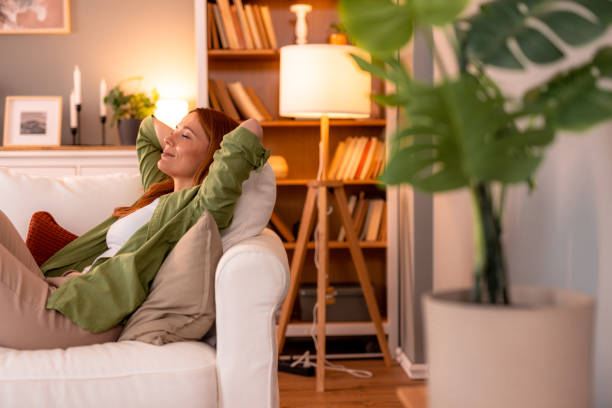 Serene woman relaxing on sofa, enjoying peaceful moment at home Redhead woman lying on a comfortable white sofa with hands behind her head, eyes closed, enjoying a serene moment of relaxation in a cozy, light-filled living room Single british person relaxing at home stock pictures, royalty-free photos & images