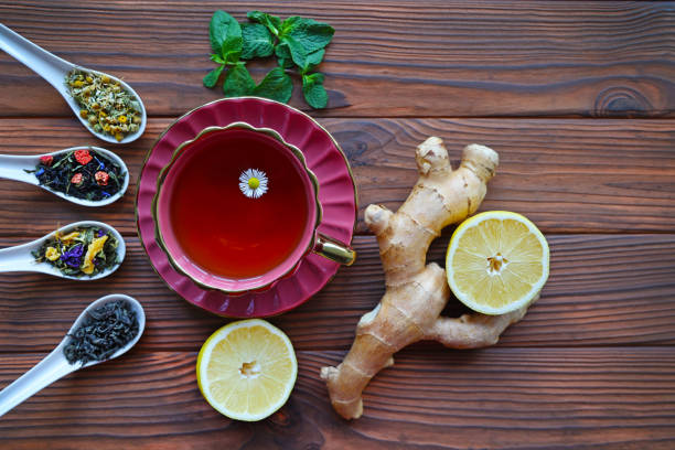 Herbal Tea with Ginger and Lemon. A vibrant cup of herbal tea surrounded by fresh ginger, lemon halves, mint leaves, and various loose tea blends on a wooden table.