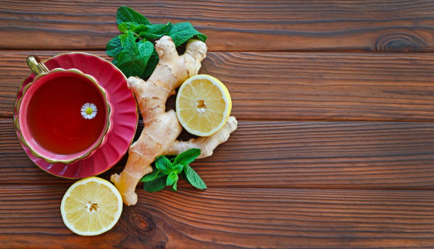 istock Herbal Tea with Ginger and Lemon Slice. A cup of herbal tea with a chamomile flower, surrounded by fresh ginger, lemon halves, and mint leaves on a wooden table. Top view. Free space for text. 2191402647