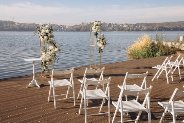 istock Wedding ceremony setup with white chairs and flower decorations on a wooden pier overlooking a lake 2191407544