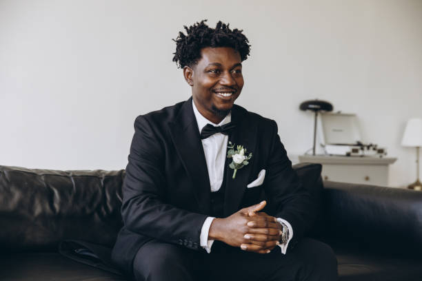 istock Groom smiling and sitting on sofa wearing black suit and bow tie 2191408485