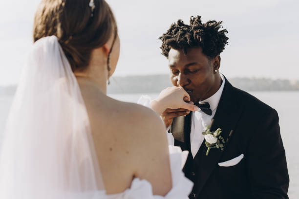 Elegant groom kissing the hand of his bride during interracial wedding ceremony by the lake