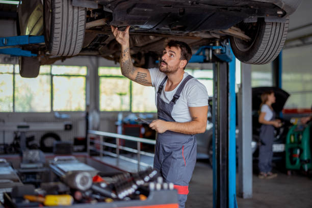 Professional Mechanic in Overalls Checking Car's Underside in Repair Shop A mechanic checks the undercarriage of a car, showcasing attention to detail and expertise in a well-lit and busy workshop environment. mechanic working under car stock pictures, royalty-free photos & images