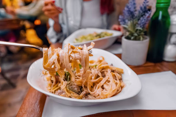 Creamy tagliatelle pasta with sauce being lifted by a fork in a restaurant, with blurred diners in the background