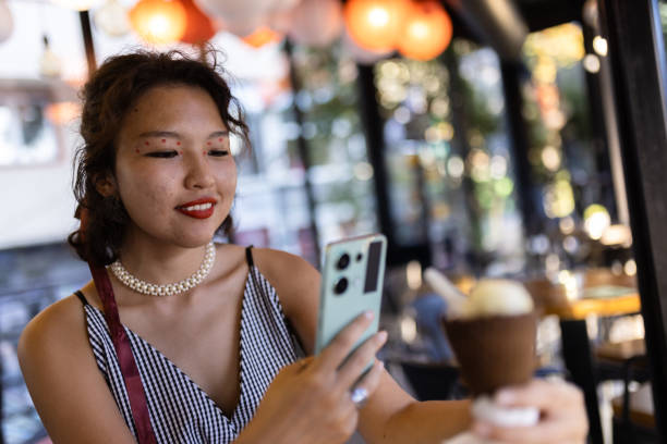 istock Young woman enjoying ice cream at a cafe while using her smartphone for taking a picture. Capturing casual moments of summer leisure with stylish makeup and fashion. 2191419755